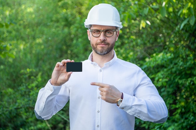 Engenheiro de homem no capacete apontando o dedo para o espaço de cópia do cartão de contato em branco ao ar livre