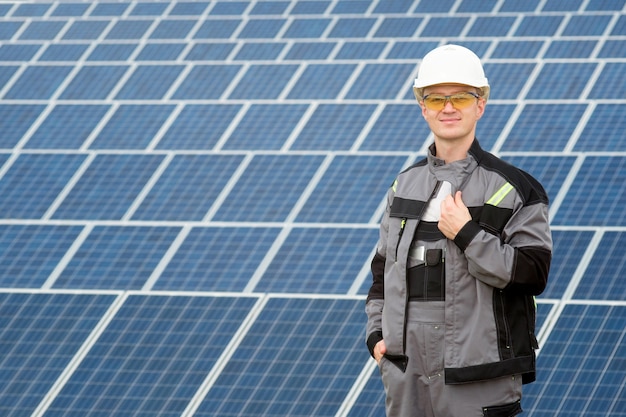 Engenheiro de painéis solares em barril branco, óculos de proteção amarelos e roupa cinza em pé perto do campo de painéis solares. conceito de energia renovável e limpa, tecnologia. copie o espaço. homem a trabalhar.