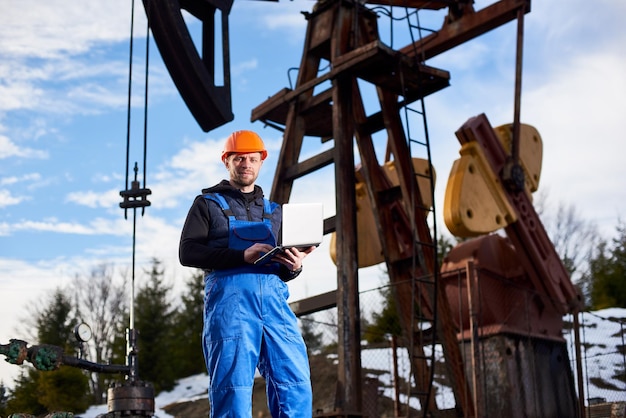 Engenheiro de petróleo com um laptop ao lado de uma plataforma de petróleo fazendo anotações em seu computador