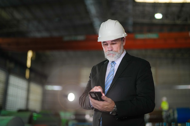 Engenheiro de retratos Chefe sênior em pé confiante e segurando smartphone na fábrica da indústria Conceito de operação inteligente da indústria e engenheiro mais velho de manutenção com bigode de barba no rosto em pé