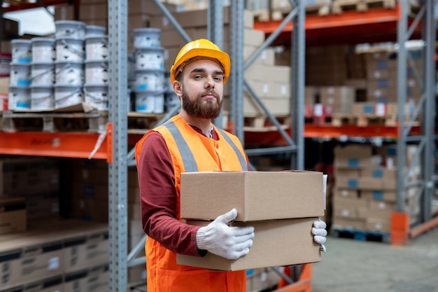 Engenheiro em roupas de trabalho segurando caixas contra prateleiras com mercadorias