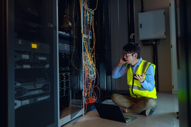 Engenheiro masculino asiático com laptop falando no smartphone enquanto inspeciona na sala do servidor do computador. conceito de gerenciamento e manutenção de servidor de banco de dados