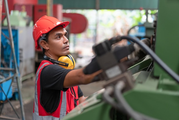 Engenheiro masculino asiático em colete de segurança vermelho e capacete verificando e reparando máquinas CNC antigas na fábrica