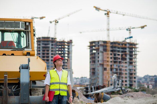 Foto engenheiro na frente de uma escavadora em um canteiro de obras com edifícios e guindastes ao fundo.