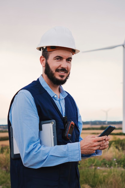Engenheiro ou técnico caucasiano de barba vertical com capacete usando um smartphone sorrindo e olhando