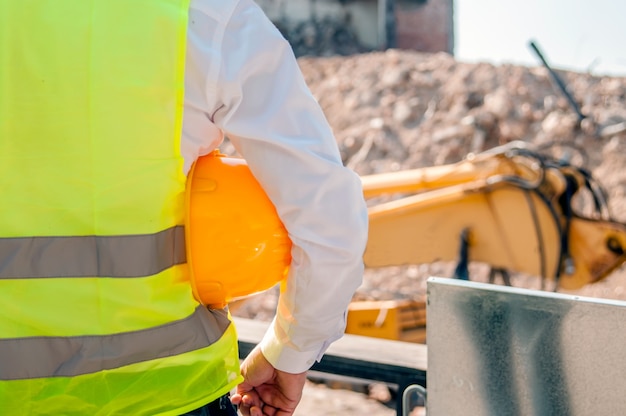 Engenheiro que segura o capacete amarelo para a segurança dos trabalhadores no fundo de novos edifícios de apartamentos highrise e guindastes de construção no fundo da noite céu nebuloso do por do sol silhueta guindaste levanta carga