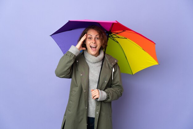 Englische Frau, die einen Regenschirm lokalisiert auf lila Raum mit Überraschungsausdruck hält