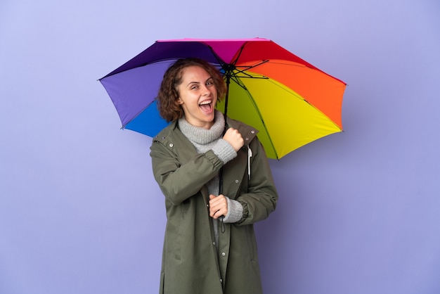 Englische Frau, die einen Regenschirm lokalisiert auf lila Wand hält, die einen Sieg feiert