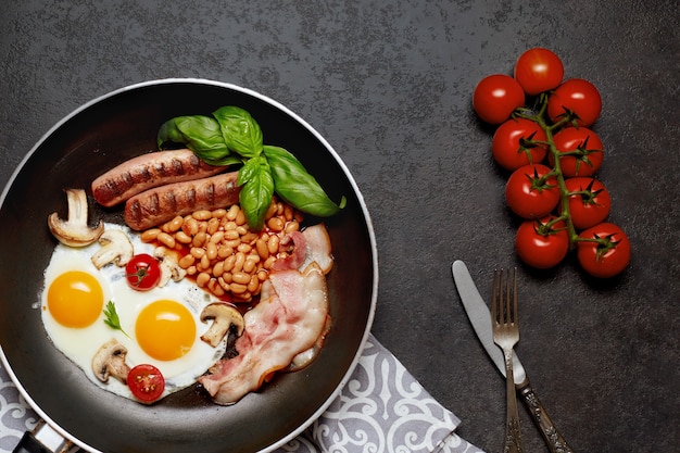 Englisches Frühstück in der Pfanne mit Spiegeleiern, Würstchen, Bohnen, Tomaten, Pilzen, Speck und Toast auf Holztisch. Speicherplatz kopieren. Draufsicht