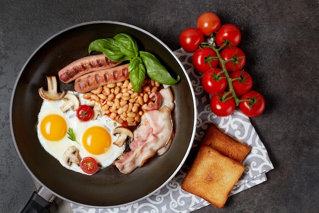 Englisches Frühstück in der Pfanne mit Spiegeleiern, Würstchen, Bohnen, Tomaten, Pilzen, Speck und Toast auf Holztisch. Speicherplatz kopieren. Draufsicht