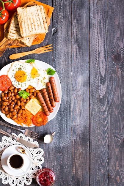 Englisches Frühstück, Spiegeleier, Würstchen, Bohnen, Toastbrot, Tomaten, Käse auf einem Holztisch,