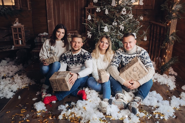 Engraçada companhia caucasiana de jovens amigos celebram o Natal juntos, sentados perto da árvore de natal em um ambiente aconchegante