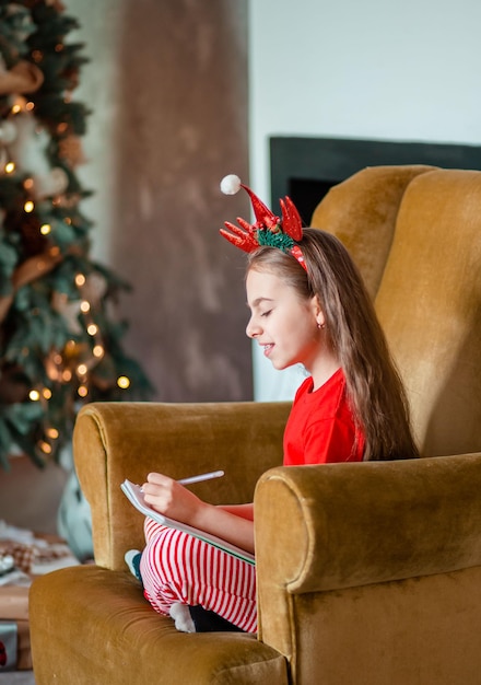 Engraçada e fofa caucasiana canhota escreve uma carta para o Papai Noel perto da árvore de Natal