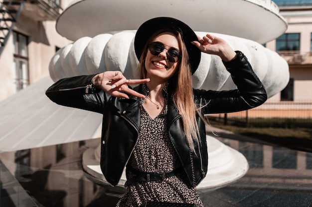 Foto engraçada e linda garota com um sorriso em um vestido da moda com uma jaqueta de couro, chapéu e óculos de sol mostra um símbolo da paz e fica na cidade