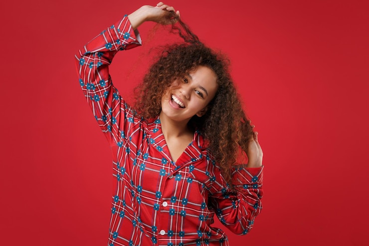 Foto engraçada jovem afro-americana em homewear de pijama posando enquanto descansava em casa isolado no retrato de estúdio de fundo vermelho. relaxe o conceito de estilo de vida de bom humor. mock up espaço de cópia. tocando o cabelo.