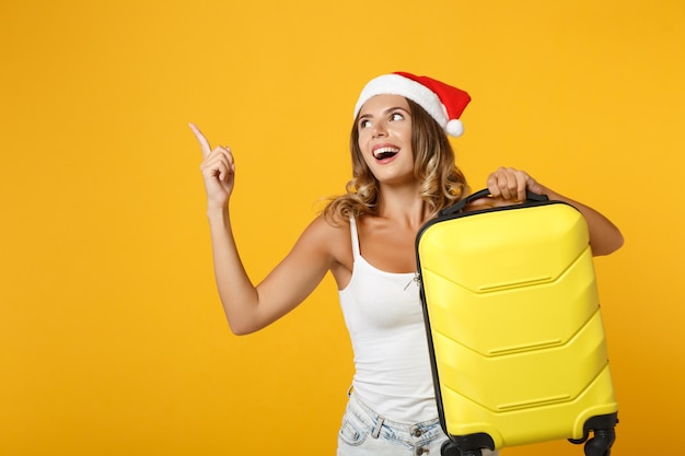 Engraçada jovem Papai Noel na camisa branca, chapéu de Natal posando isolado no fundo amarelo. Feliz Ano Novo 2020 conceito de feriado de celebração. Mock up espaço de cópia. Segure a mala, apontando o dedo indicador para cima.