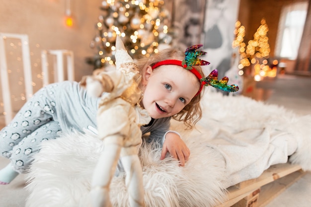 Engraçada linda menina criança com chifres de veado em um pijama da moda deitada na cama e dando um brinquedo no fundo de luzes e decorações de Natal