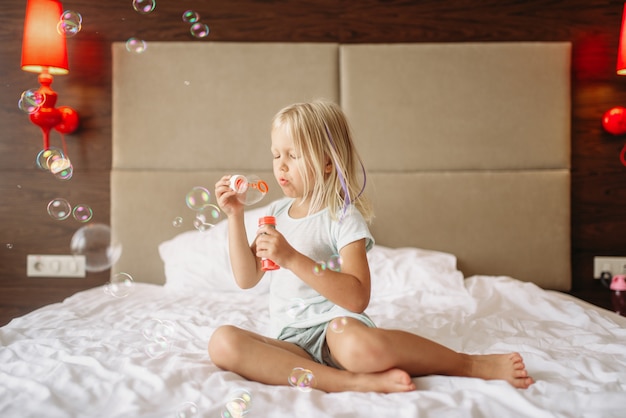 Engraçadinha menina deitada na cama e soprando bolhas. Uma infância verdadeiramente despreocupada, momentos felizes