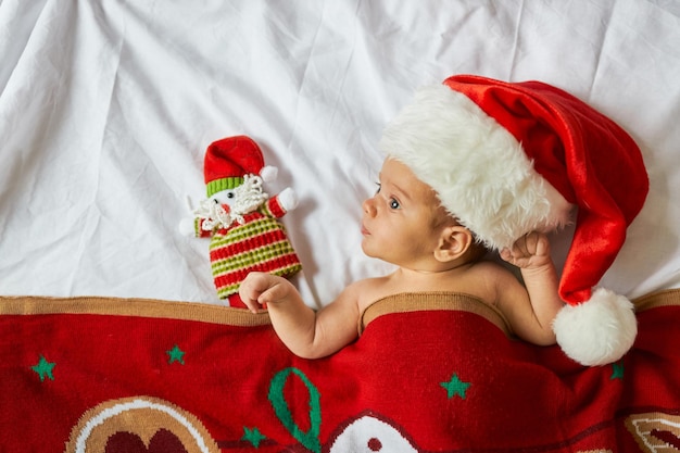 Engraçado adorável bebê recém-nascido, usando chapéu de Papai Noel, encontra-se sob o cobertor vermelho de Natal com brinquedo. Vista de cima, casa de Natal dentro de casa