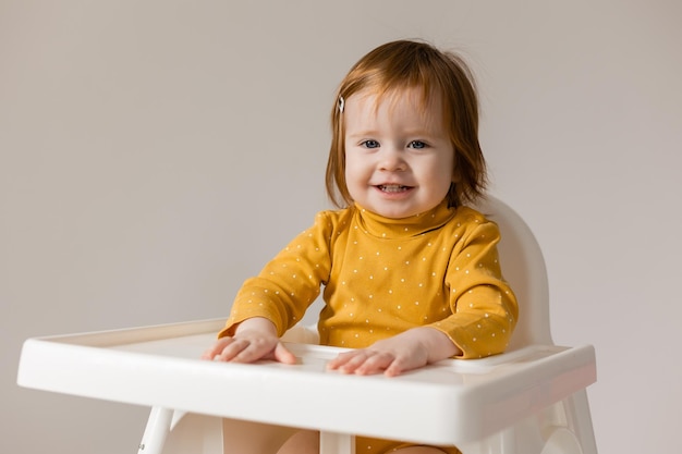 engraçado bebê ruivo de olhos azuis em um body amarelo sentado em uma cadeira alta branca