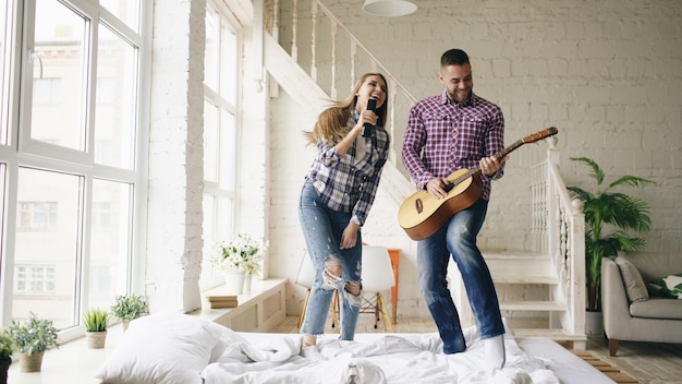 Engraçado casal feliz e amoroso dança na cama cantando com controlador de tv e tocando guitarra