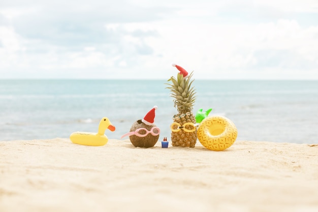 Engraçado e atraente abacaxi e coco em elegantes óculos de sol na areia contra o mar azul-turquesa. Usando chapéus de Natal.