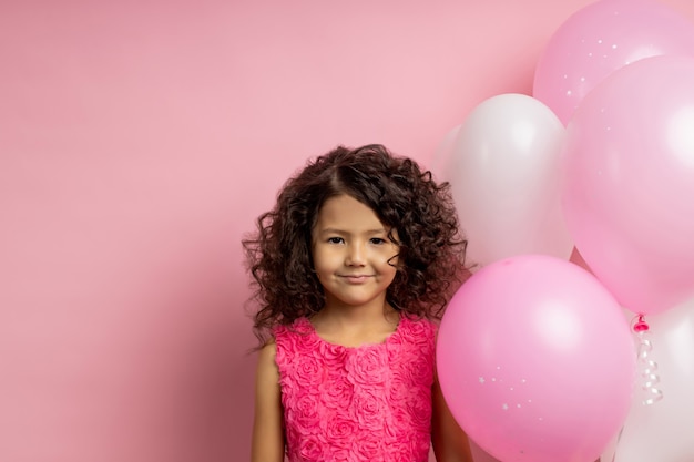 Engraçado garoto atraente com cabelo encaracolado, em pé perto de balões de ar rosa e branco na festa de aniversário, com um lindo vestido, sorrindo, isolado na parede rosada com espaço de cópia. infância feliz, férias.