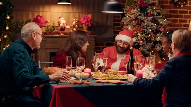 Engraçado jovem adulto disfarçado de Papai Noel discutindo com as pessoas enquanto comemora o feriado de inverno. Homem festivo vestindo fantasia de Papai Noel no jantar de Natal conversando com membros da família