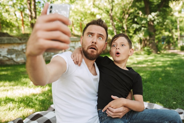 Engraçado jovem pai sentado com seu filho pequeno fazer selfie pelo telefone celular.