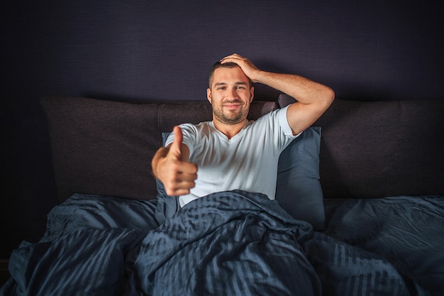 Engraçado jovem sentado na cama e sorrindo. ele mostra o polegar grande em uma mão e segura outra na cabeça. guy está satisfeito e feliz.