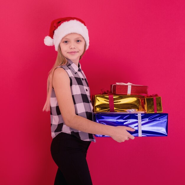 Engraçado sorridente criança alegre menina com chapéu de Papai Noel segurando o presente de Natal na mão sobre o fundo vermelho