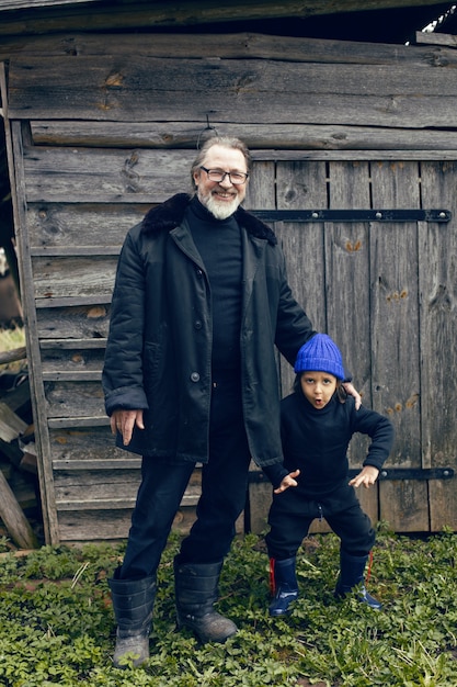 Enkel mit blauem Hut und Stiefeln mit einem Großvater in einem Schaffellmantel steht im Frühjahr in Russland in einem Holzhaus im Dorf