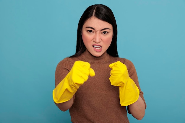 Enojada y agresiva ama de casa asiática de pie en posición de lucha en las tareas domésticas guantes de goma aislados en un fondo azul, Listo para su deber, Concepto de hogar de limpieza, Expresión facial femenina en confianza