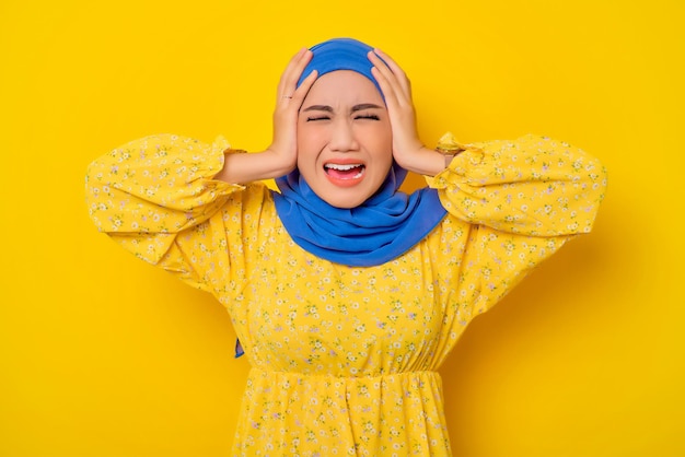 Enojada, joven y hermosa mujer asiática musulmana cubriendo los oídos con la mano y gritando con los ojos cerrados aislada sobre el fondo amarillo