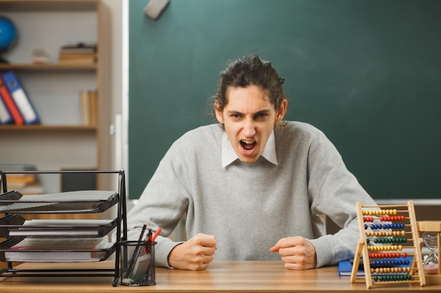 enojado mirando a la cámara joven maestro sentado en el escritorio con herramientas escolares en el aula