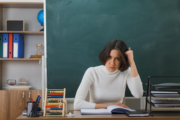 Enojado poniendo la mano en la cabeza joven maestra sentada en el escritorio con herramientas escolares en el aula