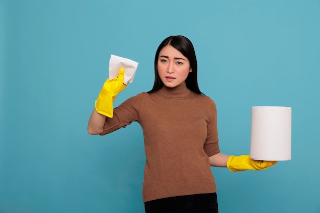 Enojado, triste y deprimido trabajador doméstico asiático sosteniendo papel tisú en el guante amarillo de pie contra un fondo azul, concepto de hogar de limpieza, mujer infeliz sintiéndose cansada frustrada y negativa