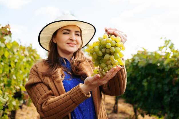 Enólogo sorrindo, mulher adulta com chapéu segurando um cacho de uva branca agricultora olhando veio ...