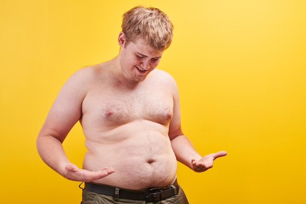 Enorme jovem engraçado com corpo nu toca barriga grande e gorda em fundo amarelo no estúdio. Conceito de obesidade, fast food e junk food, esportes, lipoaspiração, perda de peso