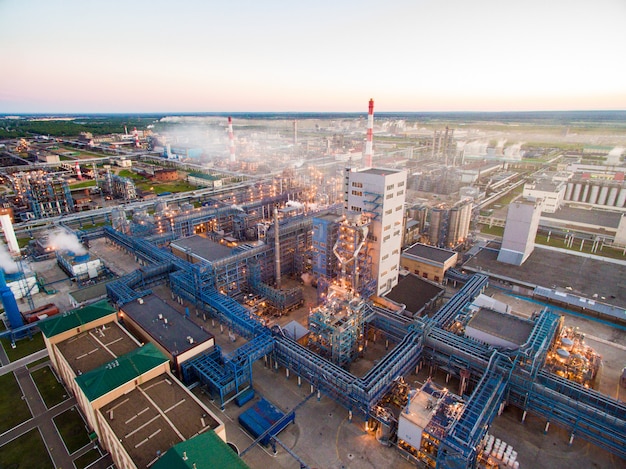 enorme refinaria de petróleo com estruturas metálicas, tubulações e destilação do complexo com luzes acesas ao entardecer. Vista aérea