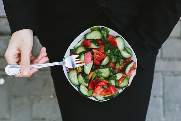 Ensalada en el almuerzo de negocios Retrato de una hermosa mujer de negocios moderna y sonriente con un traje negro con un plato de ensalada de verduras en las manos que come en la calle cerca del trabajo