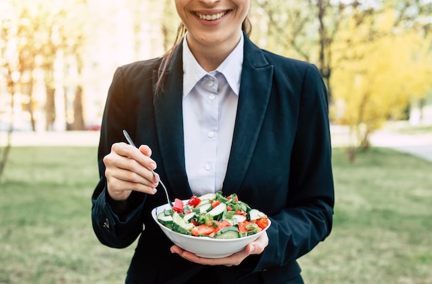 Ensalada en el almuerzo de negocios Retrato de una hermosa mujer de negocios moderna y sonriente con un traje negro con un plato de ensalada de verduras en las manos que come en la calle cerca del trabajo