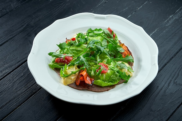 La ensalada de aperitivo tradicional italiana es vitello tonnato. Ternera en rodajas finas con rúcula, tomate y pesto, servida en un plato blanco sobre un fondo oscuro