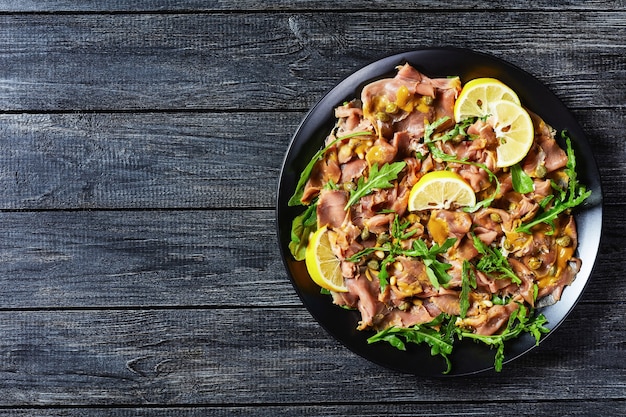 Ensalada de atún en rodajas finas con alcaparras, rúcula, piñones y aderezo de mostaza en un plato negro sobre una mesa de madera, vista horizontal desde arriba, plano, espacio libre