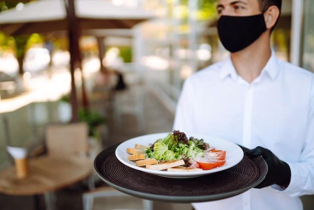 Ensalada en la bandeja de un camarero Joven camarero con mascarilla protectora mientras trabaja en un café reabierto