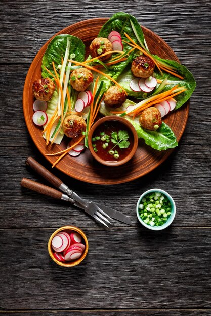 Ensalada banh mi vietnamita con albóndigas de cerdo zanahoria pepino y rábano servido en ensalada de lechuga romana en un plato marrón con salsa de chile dulce sobre fondo de madera rústica oscura vista superior primer plano