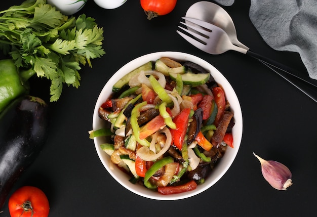 Ensalada de berenjenas, tomates, pimientos y pepinos en un tazón blanco sobre fondo negro