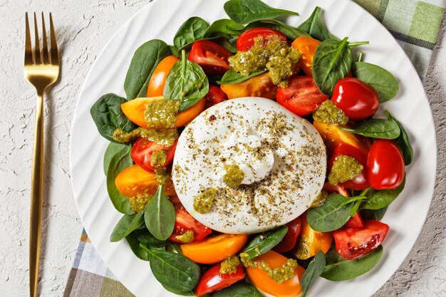 Ensalada Burrata Caprese con espinacas y salsa de pesto de albahaca sobre un plato blanco sobre una mesa de hormigón, vista horizontal desde arriba, puesta plana, espacio libre