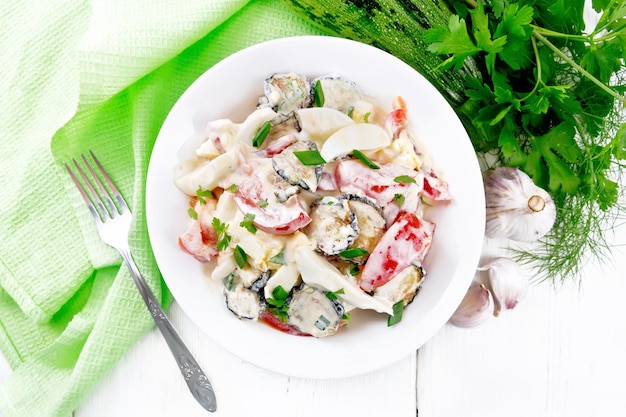 Ensalada de calabacín frito, huevo duro, tomate fresco y ajo, sazonado con mayonesa o crema agria en un plato, servilleta y perejil sobre fondo de tablero de madera desde arriba