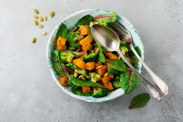 Ensalada con calabaza al horno, acelgas, brócoli y pipas de calabaza en plato de cerámica sobre mesa de piedra u hormigón. Estilo rústico.